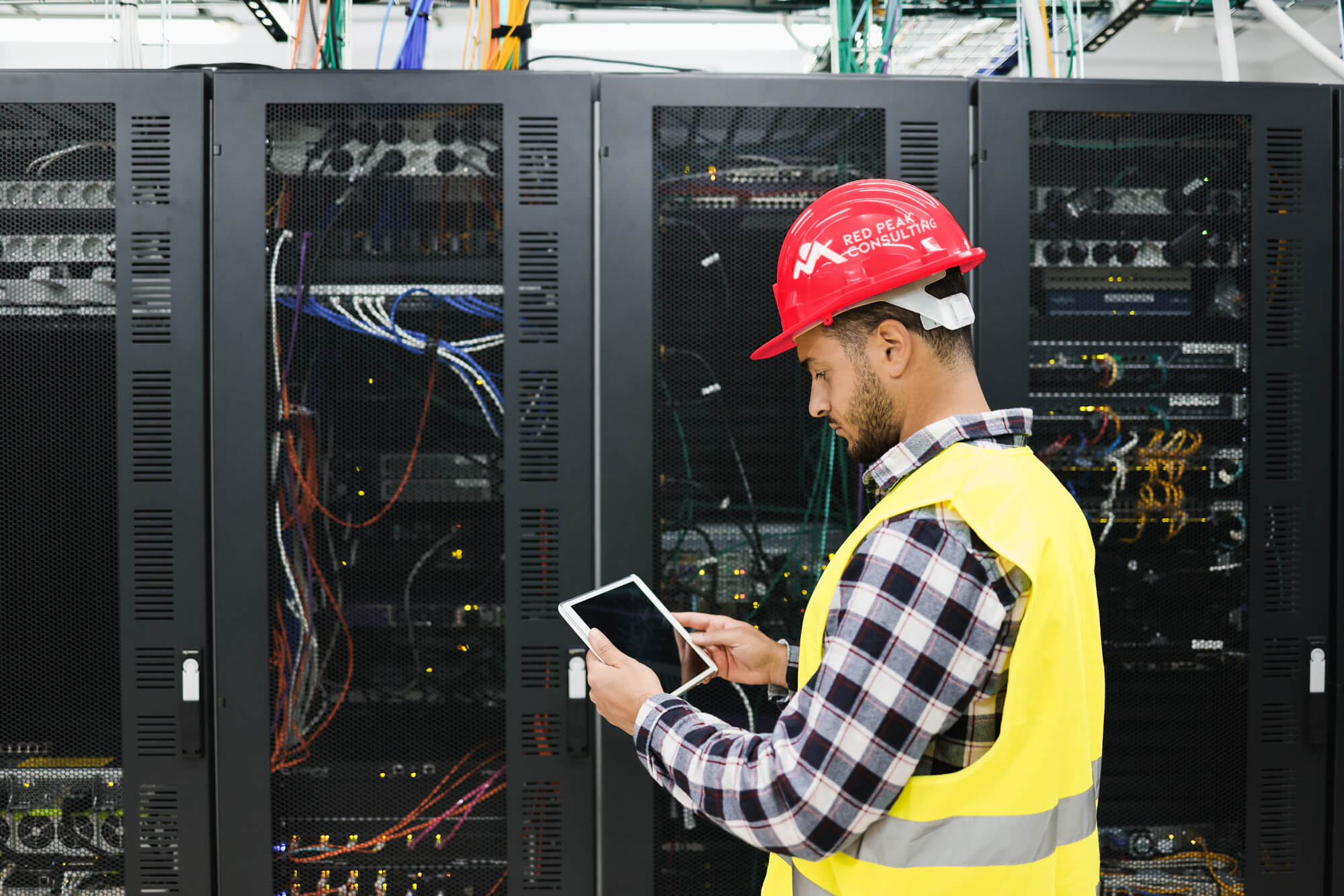 Worker in a hard hat using a tablet to monitor servers in a data centre.