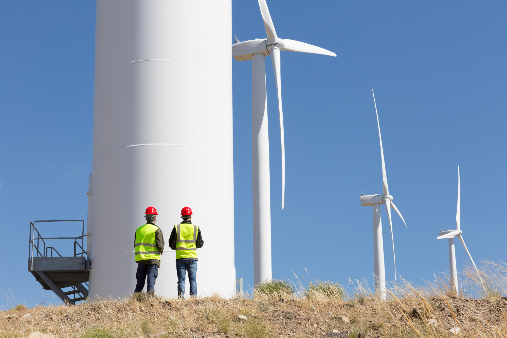 Two workers wearing hard hats standing at the base of a wind turbine, inspecting and coordinating operations.