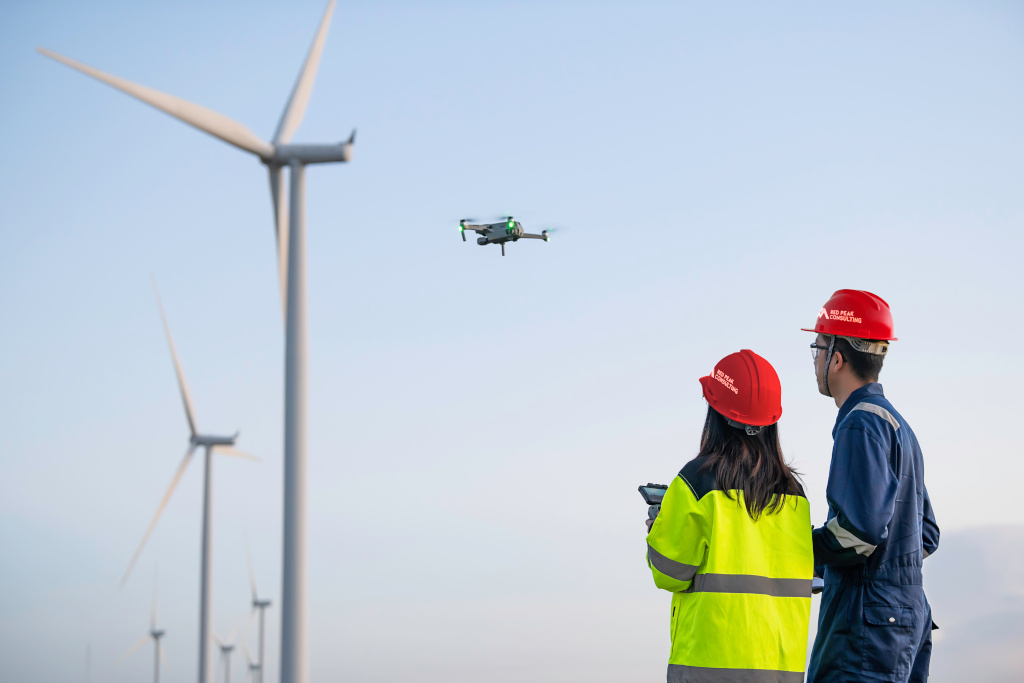 Two workers wearing hard hats operating a drone near a wind turbine for inspection, illustrating delivery and project controls.